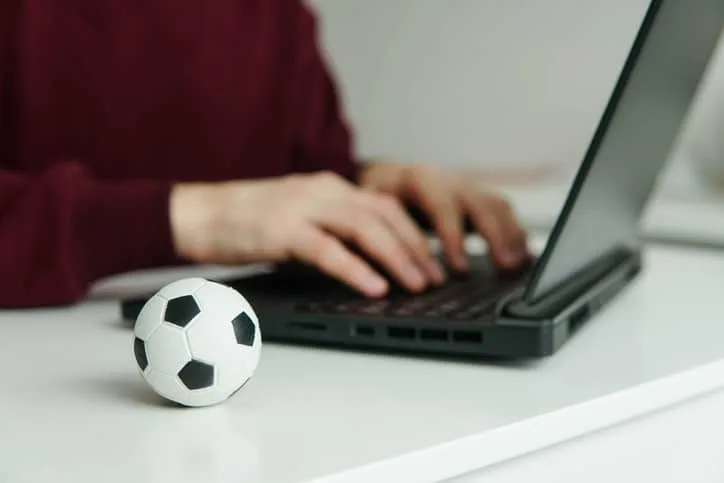 Jovem lendo notícias de futebol ou escrevendo artigo de futebol no laptop. Bola de futebol sobre a mesa. Apostas, conceito de jogo