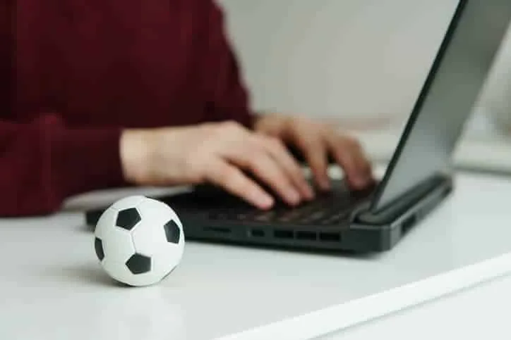 Jovem lendo notícias de futebol ou escrevendo artigo de futebol no laptop. Bola de futebol sobre a mesa. Apostas, conceito de jogo.