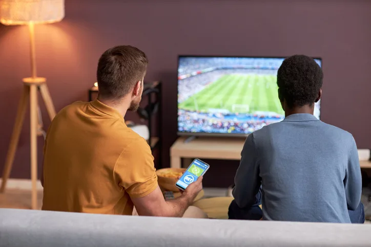 Duas silhuetas de homens assistindo a uma partida de futebol na TV, cada um segurando um smartphone com uma plataforma de apostas online aberta.